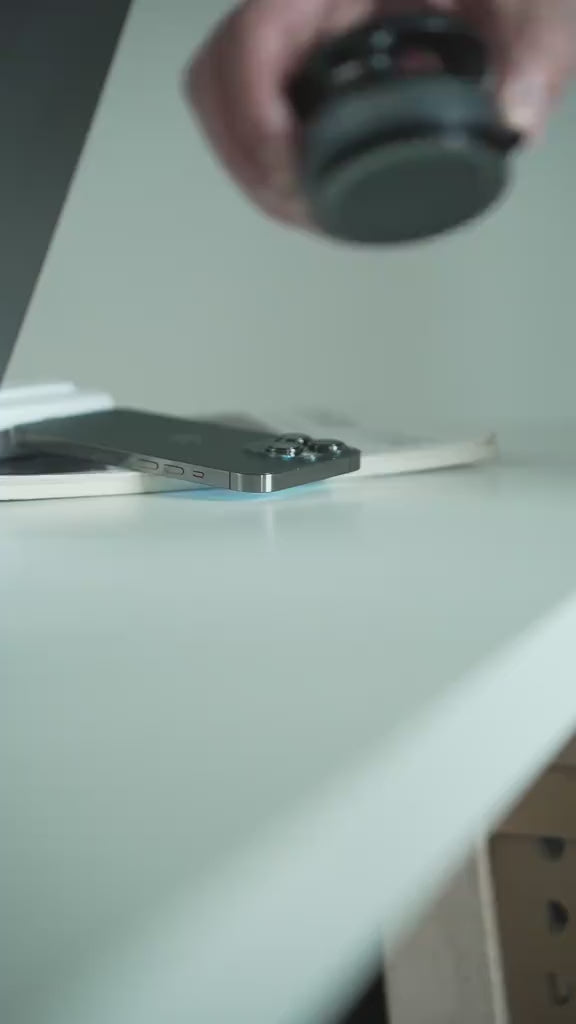 Close-up of a black magnetic car phone holder being placed on a white desk with a smartphone and a tablet nearby, showcasing its sleek design and magnetic grip.